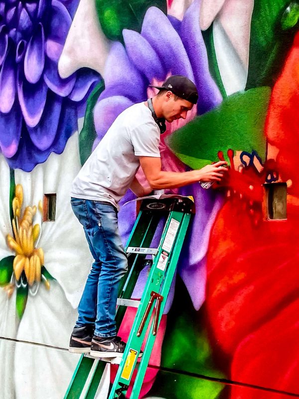 Artist Brandon Marshall paints a large, colorful mural while standing on a ladder.