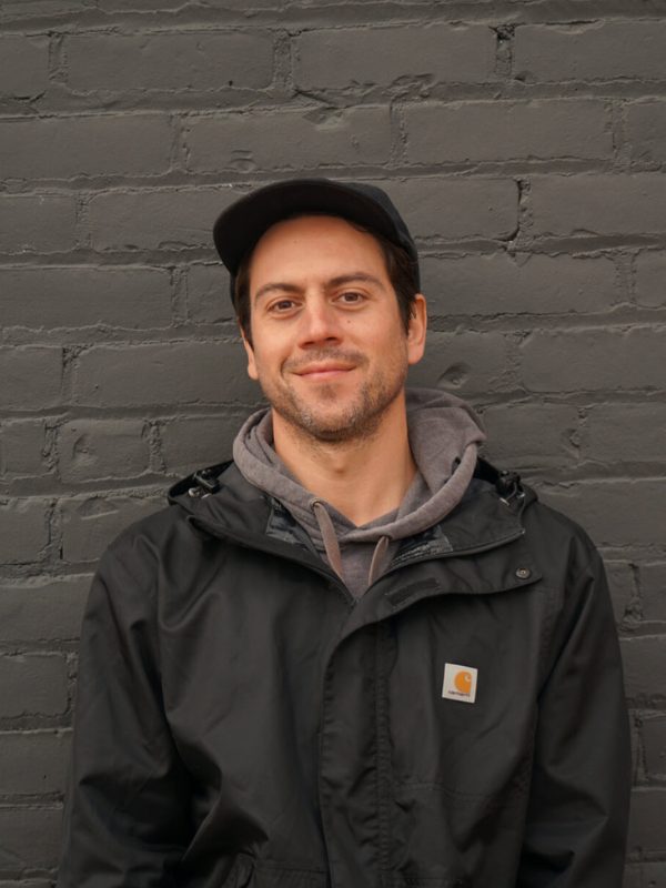 Artist Jesse Kassel smiling in front of a grey brick background.